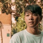 Portrait of an Asian man with short black hair in a sunlit urban setting.
